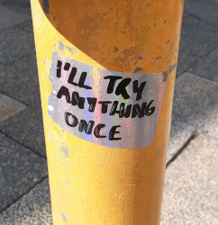 A shiny silver sticker on a traffic light pole with the text 'I'll Try Anything Once' written on it in black sharpie.